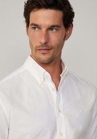 Camicia bianca in cotone con colletto a punta, caratterizzata da ricami sottili e accenti interni di un blu chiaro. Tessuto liscio e fresco.