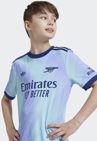 Maillot de football bleu clair en tissu lisse, avec des accents bleu foncé, le logo Emirates et un emblème d'équipe stylisé.