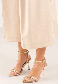 Sandalias de tacón alto con tiras en dorado metálico, que presentan una punta afilada y una correa al tobillo. Detalladas con un pequeño acento decorativo y un tacón elegante.