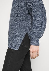 Mörkblå stickad tröja med ett texturerat mönster, sprund i sidsömmarna och ribbade muddar, ihop med svarta jeans.