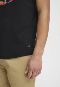 Sort bomulds t-shirt med korte ærmer, prydet med farverigt grafisk design foran. Vævet label nær kanten. Tan cargo shorts vist.
