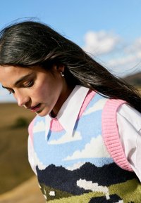 Frau mit langen dunklen Haaren, die silberne Ohrringe und eine bunte Strickweste über einem weißen Hemd mit Kragen trägt, im Freien unter einem blauen Himmel mit Wolken.
