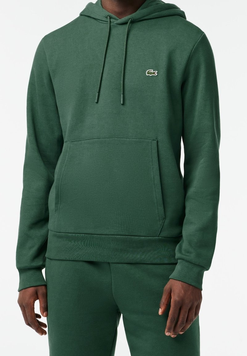 Homme portant un sweat à capuche vert foncé et un pantalon de jogging assorti, avec un petit logo crocodile sur la poitrine, mains détendues sur les côtés.