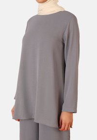 Long-sleeve tunic in textured gray fabric, featuring a loose fit and rounded hem, paired with a beige scarf around the neck.