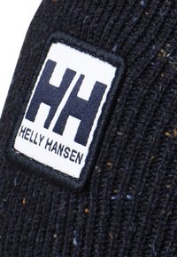Gebreide beanie van donkerblauwe garen met een textuurpatroon; heeft een witte patch met "HH" en "HELly HANSEN" in vetgedrukte zwarte letters.
