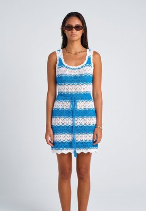 Donna in piedi che indossa un abito senza maniche a righe blu e bianche lavorato a maglia con fiocco in vita e occhiali da sole scuri su sfondo chiaro uniforme.