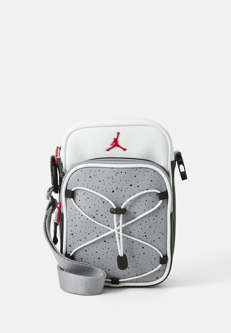 Šedobílá crossbody taška s tečkovaným designem, zobrazující červené logo, se zipovým uzavíráním a nastavitelným popruhem. Obsahuje bungee detail na přední straně.