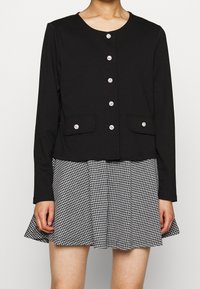 Černý zkrácený kardigan s kulatým výstřihem, zdobený stříbrnými knoflíky a dvěma předními kapsami, kombinovaný s plisovanou sukní v černobílé kostce.