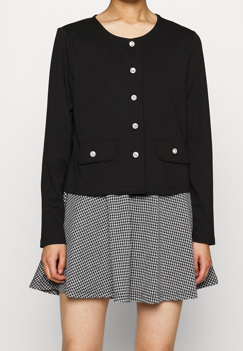 Černý zkrácený kardigan s kulatým výstřihem, zdobený stříbrnými knoflíky a dvěma předními kapsami, kombinovaný s plisovanou sukní v černobílé kostce.