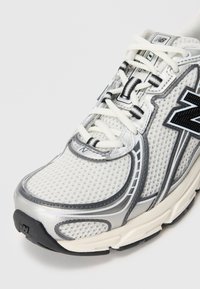 Baskets de course New Balance blanches et argentées avec un dessus en mesh, logo noir et lacets blancs sur un fond clair.