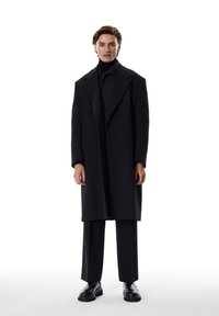 Abrigo de lana negro con grandes solapas, combinado con un cuello alto y pantalones a juego. Textura suave y ajuste a medida. Zapatos negros completan el look.