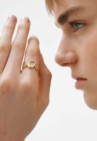 Anillo de oro con acentos circulares y decorativos, llevado en una mano. Primer plano de dedos y perfil de una persona desenfocado.
