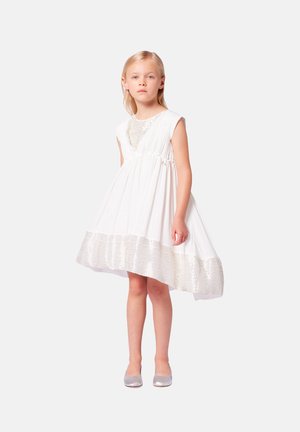Robe blanche avec des manches courtes, un corsage texturé et une jupe évasée avec un ourlet à motifs. Une paire de ballerines argentées.