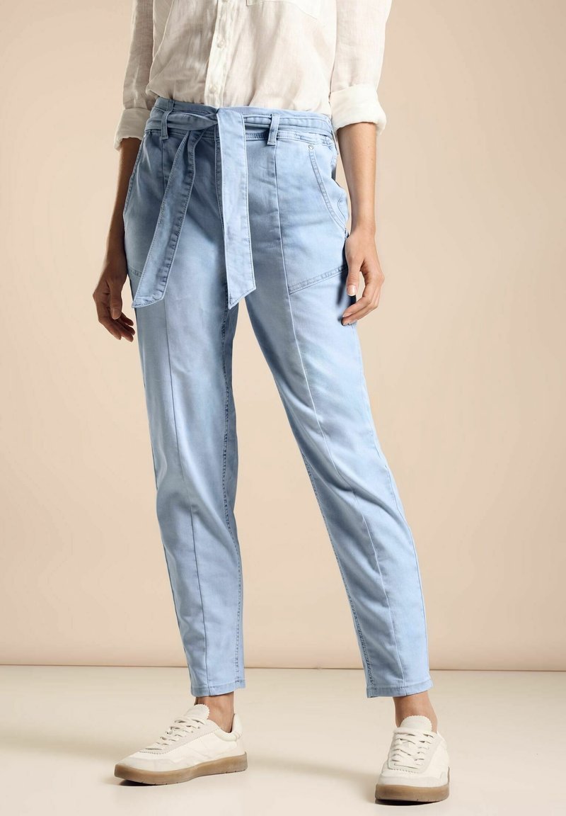 Pantalones azul claro con cinturón en tejido suave y piernas ajustadas. Presentan bolsillos frontales y un diseño informal, combinados con zapatillas blancas.