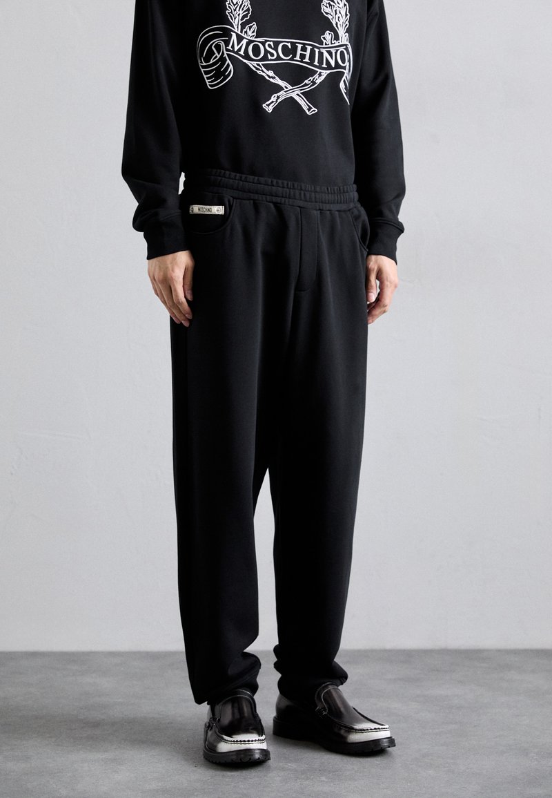 Svarta sweatpants med en avslappnad passform, elastisk midja, fickor och varumärkesdetaljer. Bärs med blanka svarta skor med silverdetaljer.