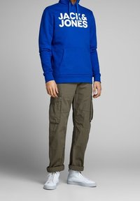 Uomo che indossa un hoodie blu di Jack & Jones, pantaloni cargo verde oliva e sneaker alte bianche, in piedi davanti a uno sfondo grigio.
