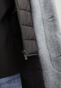 Tela exterior de mezcla de lana gris con un forro acolchado negro. Cuenta con una cremallera y un diseño de bolsillo minimalista. Superficies texturizadas con colores en contraste.