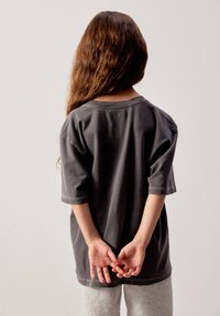 Enfant aux longs cheveux bruns, debout de dos, portant un t-shirt sombre surdimensionné et un pantalon gris clair, les mains jointes derrière le dos.