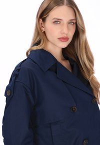 Marineblauwe trenchcoat met een dubbele rij knopen, grote revers en knoopdetails. Gladde textuur met een nauwsluitende pasvorm.