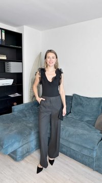 Svart body med volangermet armer, kombinert med høytlivede grå bukser. Modellen har på seg svarte spisse hæler og står på et teksturert gulv.