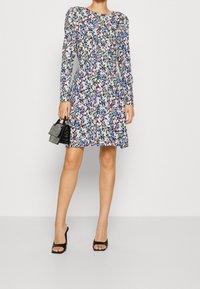 Długa sukienka z długim rękawem w czarnym kolorze z wielokolorowymi kwiatami, z taliowaniem, rozkloszowanym dołem, zestawiona z małą czarną torebką i butami na obcasie odkryte palce.