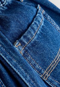 Gros plan sur un tissu en denim bleu montrant des coutures détaillées et une poche arrière sur un jean.