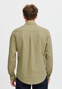 Cămașă lungă de culoare verde măslin, cu guler cu butoane, plisă la spate și hem rotunjit. Țesătură moale cu textură fină și mansete cu butoane.