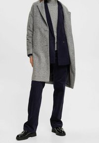 Manteau en laine gris avec un design long et structuré, superposé à un pull en maille grise texturée et un pantalon en velours côtelé bleu foncé, complété par des chaussures noires.
