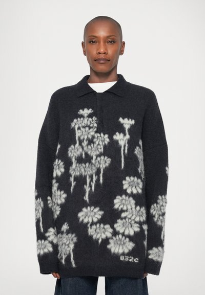 Personne portant un pull oversized foncé avec des motifs floraux abstraits blancs, un col et des boutons, associé à un jean bleu.