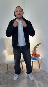 Μαύρο φλις μπουφάν με φερμουάρ πάνω από λευκό πουκάμισο, μαύρα τζιν και λευκά αθλητικά παπούτσια. Στέκεται σε ένα φωτεινό δωμάτιο με υφή στον τοίχο και μια καρέκλα.