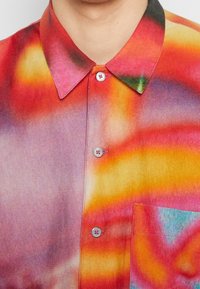 Chemise à manches courtes colorée avec un motif en dégradé dans des tons rose, orange et vert. Elle possède un col pointu et des boutons décoratifs.