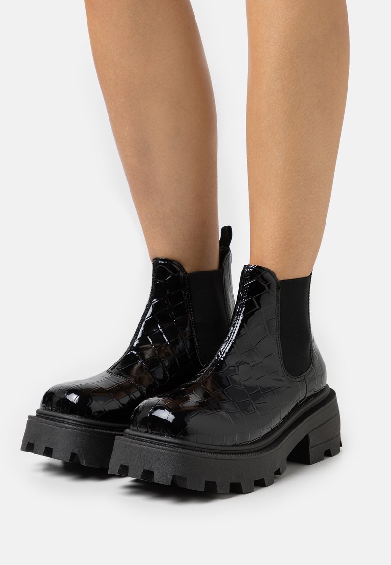 Bottines noires brillantes à semelles épaisses à crampons et texture crocodile portées sur des jambes nues sur fond blanc.