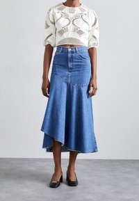 Top court blanc avec des motifs texturés, associé à une jupe en denim bleu à ourlet asymétrique et bords effilochés. Chaussures à talons blocs noires.