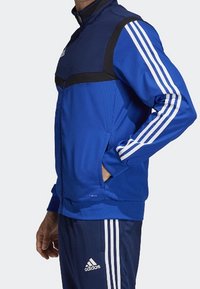 Blå träningsjacka med svarta detaljer, dragkedja vid kragen och vita tredubbla ränder på ärmarna; tillverkad av lätt och strukturerad tyg.