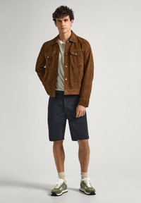 Chaqueta de ante marrón, camiseta de rayas, pantalones cortos oscuros y zapatillas verdes con detalles en blanco. La persona está de pie en un fondo neutral.