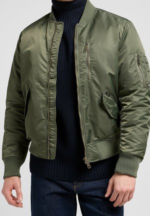 Homme portant une veste bomber vert olive avec poches zippées et à rabat sur un pull à col roulé côtelé bleu marine et un jean bleu.