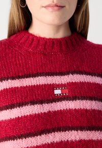 Rood en roze gestreepte gebreide trui met een textuurafwerking en een ronde hals. Heeft een klein "TOMMY JEANS" label op de voorkant.