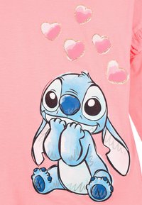 Różowa koszula z materiału z postacią z kreskówki o dużych oczach i niebieskim futrze, siedzącą z założonymi łapami, otoczoną błyszczącymi różowymi sercami.