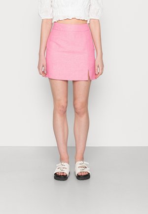Femme portant un chemisier court ajouré blanc, une mini-jupe rose avec une fente latérale, et des sandales blanches à boucle, se tenant devant un fond clair uni.