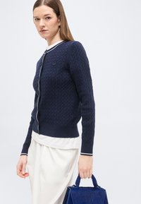 Γυναίκα με μπλε σκούρο πλεκτό ζακέτα και άσπρη φούστα, κρατώντας μια μπλε υφασμάτινη τσάντα, σε απλό ανοιχτό φόντο.