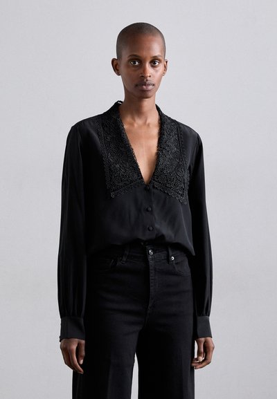 Blouse en soie noire avec un décolleté en V profond et un détail de col en dentelle complexe, dotée de manches longues et d'une fermeture à boutons, associée à un pantalon noir.