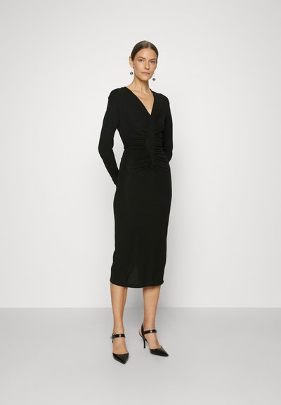 Μαύρο φόρεμα μακρύ μανίκι με βαθύ V-ντεκολτέ και συγκεντρωμένη λεπτομέρεια μπροστά. Στενή σιλουέτα, μέχρι το γόνατο, συνδυασμένο με μαύρα τακούνια.