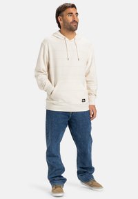 Lys beige hættetrøje med horisontale striber, justerbar hætte, frontlomme, parret med løse blå jeans og beige sneakers.