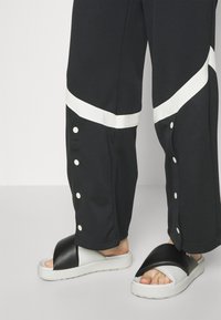 Person med svarta byxor med vita diagonala ränder och sidoknappar, tillsammans med svarta och vita slidesandaler på ett vitt golv.