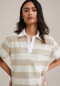 Pruhované polo tričko v béžové a bílé barvě, s krátkým rukávem, bílým límečkem a volným střihem. Měkká bavlněná textura.