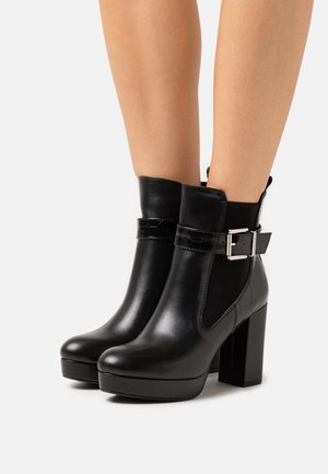Bottes cheville en cuir noir avec talon épais, texture lisse, et une sangle ornée d'une boucle argentée. Design avec panneau élastique latéral.