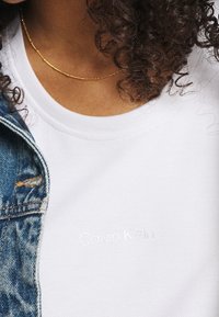 Biała bawełniana koszulka z wytłoczonym logo "Calvin Klein"; połączona z niebieską dżinsową kurtką. Złota naszyjnik z koralików dodaje szczegółów.