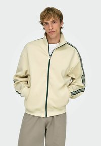 Jeune homme portant une veste beige à fermeture éclair avec des bandes noires sur les manches et un pantalon gris clair, debout les mains dans les poches.