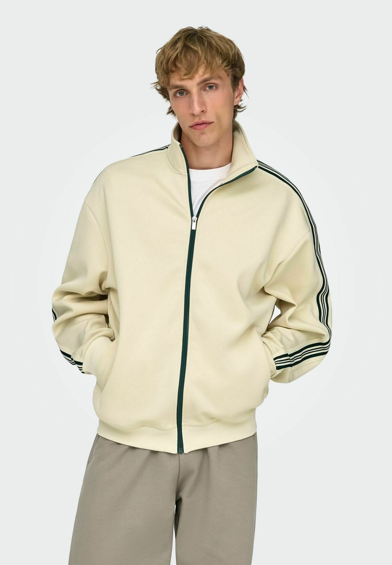 Jeune homme portant une veste beige à fermeture éclair avec des bandes noires sur les manches et un pantalon gris clair, debout les mains dans les poches.