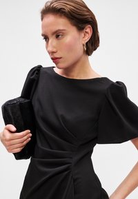 Melnā kleita ar pūkainām piedurknēm un sānu rušinājumu, izgatavota no gludas auduma. Modelis tur melnu mākslīgās kažokādas clutch somu. Vienkāršs dekoltē.